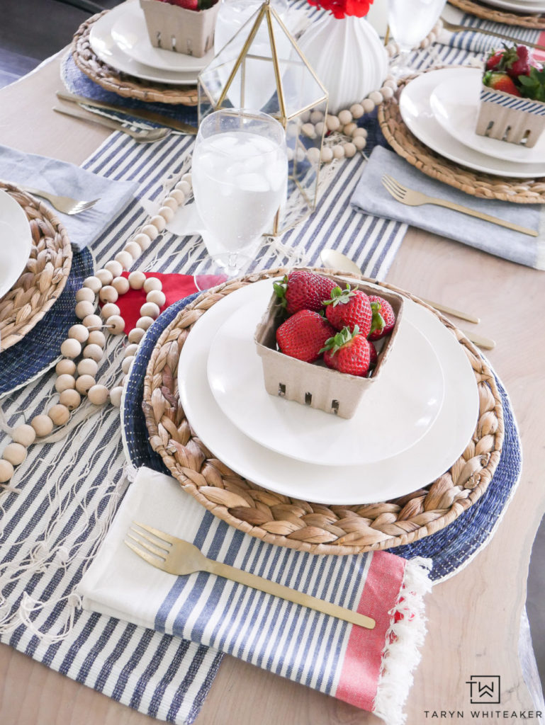 DIY Memorial Day Table Decorations featured by top AL home and lifestyle blogger, She Gave It A Go |Memorial Day Table Top Decorations by popular Alabama DIY blog, She Gave It A Go: image of a table decorated with a blue and white stripe table runner, wooden bead garland, red white and blue tassel cloth napkins, gold forks, round blue fabric placemats, sea grass chargers, two white plates, and a cardboard berry basket filled with strawberries. 