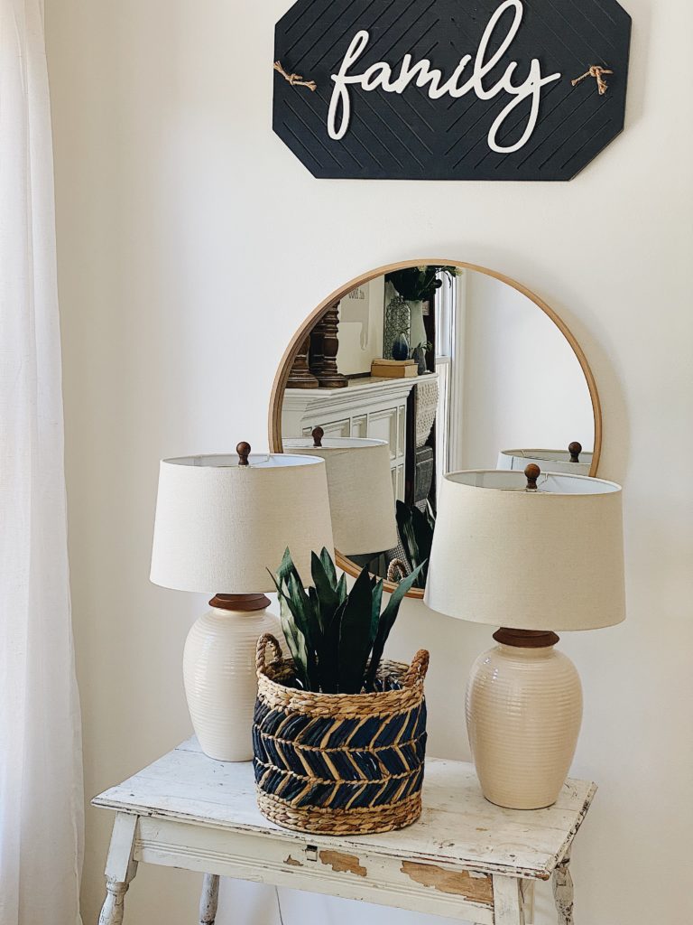 Farmhouse Fireplace Decor Ideas for Summer featured by top AL home blogger, She Gave It A Go | Farmhouse Fireplace Decor Ideas by popular AL life and style blog, She Gave It A Go: image of a white distressed table resting under a round mirror and black family sign and decorated with two white base lamps and a snake tongue plant in a woven seagrass basket. 