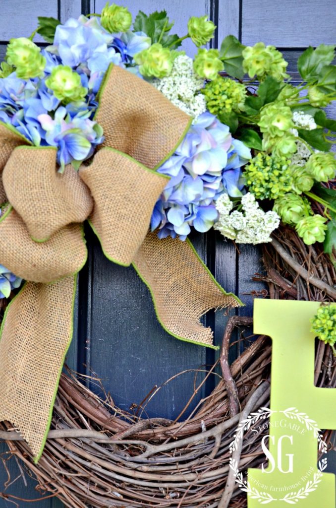 7 Rustic Farmhouse Wreath Ideas for your Summer Front Porch featured by top AL home blogger, She Gave It A Go | Farmhouse Wreath Ideas by popular Alabama life and style blog, She Gave It A Go: image of a blue green and white hydrangea wreath with a burlap ribbon and green wooden letter H. 