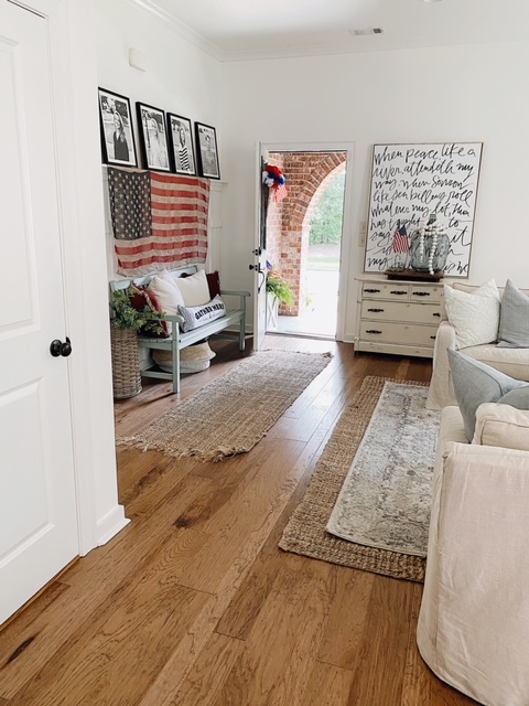Patriotic Farmhouse Decor featured by top AL home blogger, She Gave It A Go | Patriotic Decor by popular Alabama life and style blog, She Gave It A Go: image of a living room with a distressed white dresser, black and white photos in black frames, blue wooden bench with red and white throw pillows, woven runner rug and area rug, white couch and arm chair, and woven American flag hanging on a wall. 