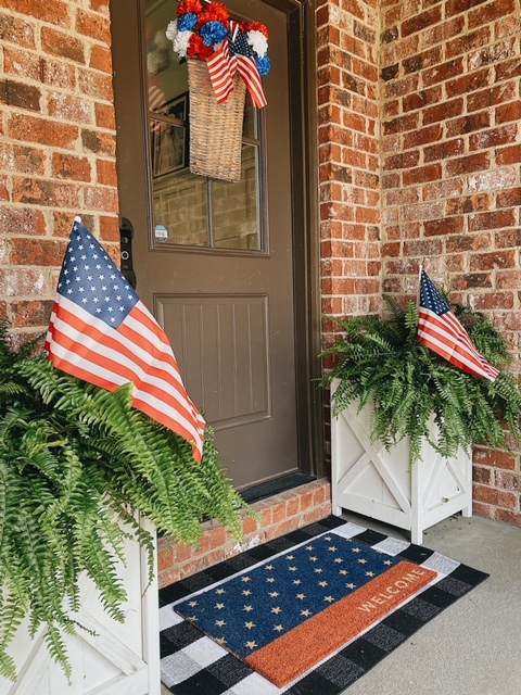 Patriotic Farmhouse Decor featured by top AL home blogger, She Gave It A Go | Patriotic Decor by popular Alabama life and style blog, She Gave It A Go: image of a front porch decorated with American flag welcome mat, white planter boxes with ferns and American flag decor, and a patriotic basket wreath filled with mini American flags and faux red, white, and blue flowers. 