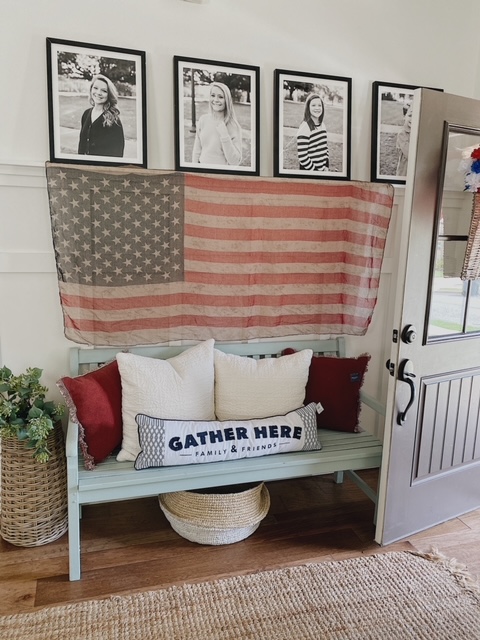 Patriotic Farmhouse Decor featured by top AL home blogger, She Gave It A Go | Patriotic Decor by popular Alabama life and style blog, She Gave It A Go: image of a living room with a distressed white dresser, black and white photos in black frames, blue wooden bench with red and white throw pillows, woven runner rug and area rug, white couch and arm chair, and woven American flag hanging on a wall. 