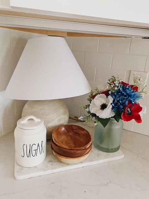 Patriotic Farmhouse Decor featured by top AL home blogger, She Gave It A Go | Patriotic Decor by popular Alabama life and style blog, She Gave It A Go: image of faux red, white, and blue flowers in a frosted glass vase on marble cutting board next to some stacked wooden bowls, sugar jar, and grey cement base lamp with a white shade. 