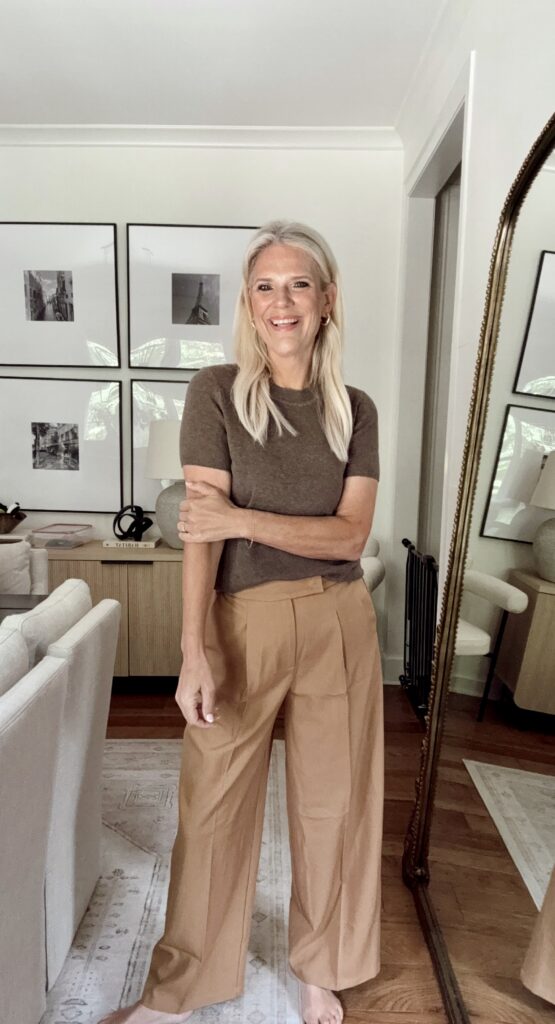Brendt Blanks wearing a brown short sleeve knit top paired with high waisted camel wide leg trousers, standing in a neutral living room with framed wall art and a floor mirror, showcasing a polished and affordable fall outfit.